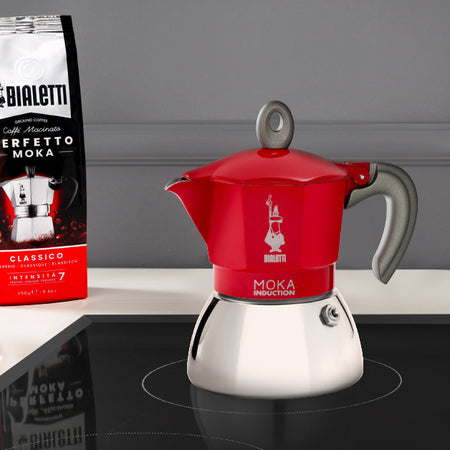 Rød Bialetti Moka Induktion 6 kopper espressokande placeret på induktionskomfur ved siden af Bialetti kaffepose.
