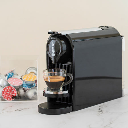Bialetti Gioia kapselmaskine brygger espresso i et glas i et køkken, kapsler i gennemsigtig beholder ved siden af.
